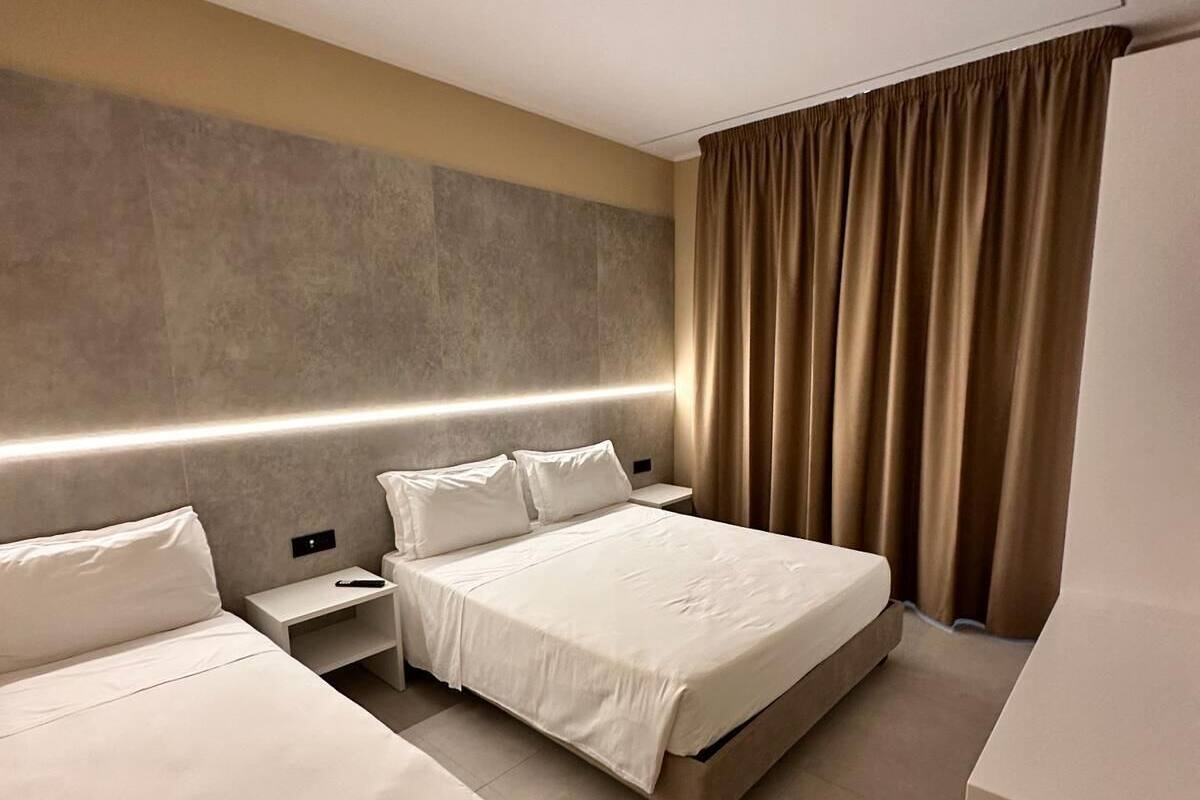 A hotel room with two beds, a bedside table, a nightstand, a large mirror, and a window with curtains.