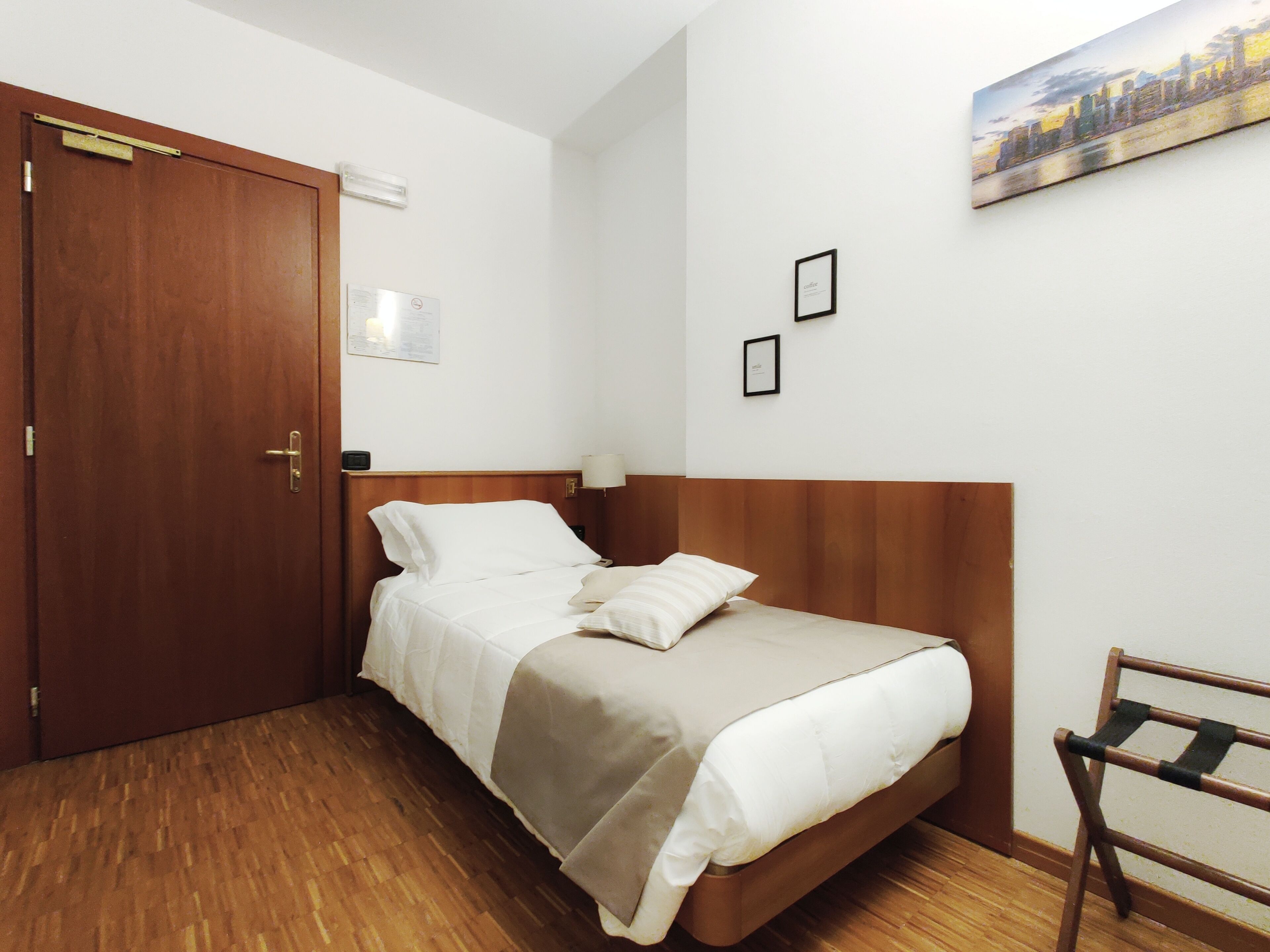 A hotel room with a wooden door, a bed with white bedding, a wooden headboard, a nightstand with a lamp, and a painting on the wall.