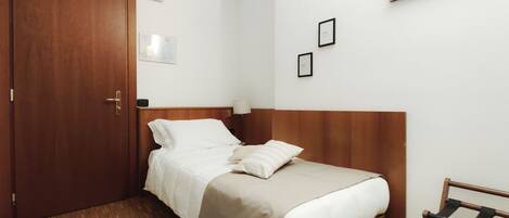 A hotel room with a wooden door, a bed with white bedding, a wooden headboard, a nightstand with a lamp, and a painting on the wall.