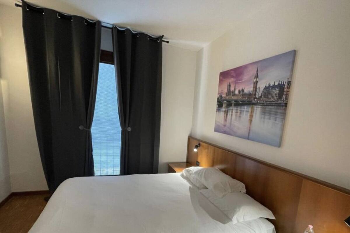 A hotel room with a bed, a nightstand, a lamp, a painting of a cityscape, and a window with curtains.