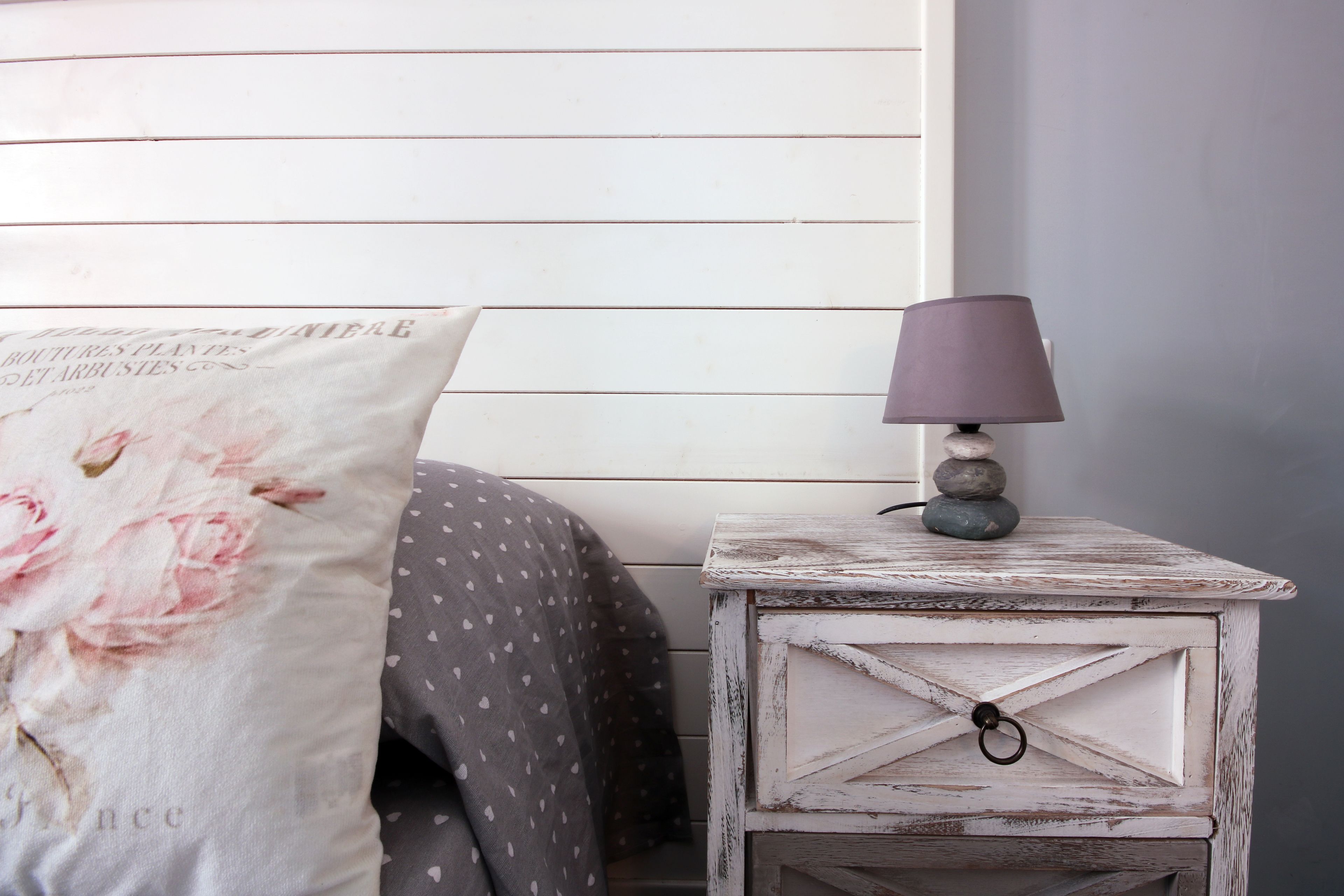 A bedroom with a bed, a white pillow with floral design, a bedside table with a lamp, and a wall with horizontal wooden slats.