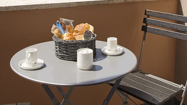 Taula rodona amb una cistada de pastissos, dues tasses blanques i una cadira a un balcó amb vistes a arbres i edificis.