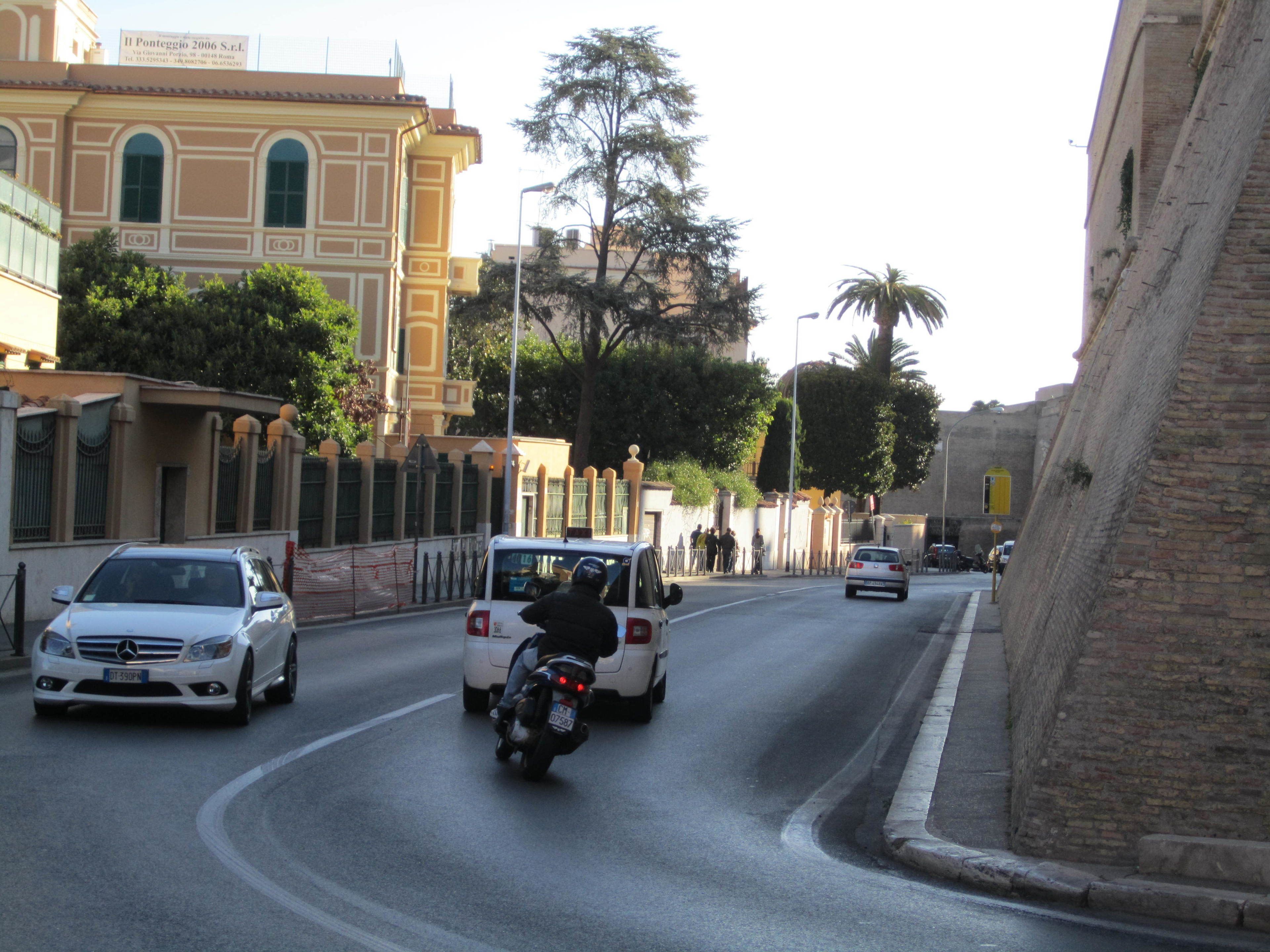 A street scene with a white car, a motorcycle, and a white van. There are buildings on both sides of the street, including a building with a yellow facade and a building with a beige facade.