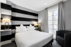 Una habitación de hotel moderna con una cama grande, una mesita de noche con lámpara, una silla y una ventana con cortinas.