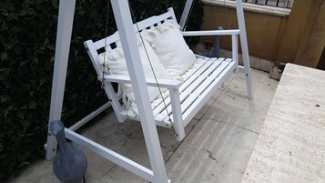 Una poltrona a dondolo bianca con cuscino, posizionata all'aperto su un pavimento in cemento con un siepe sullo sfondo.