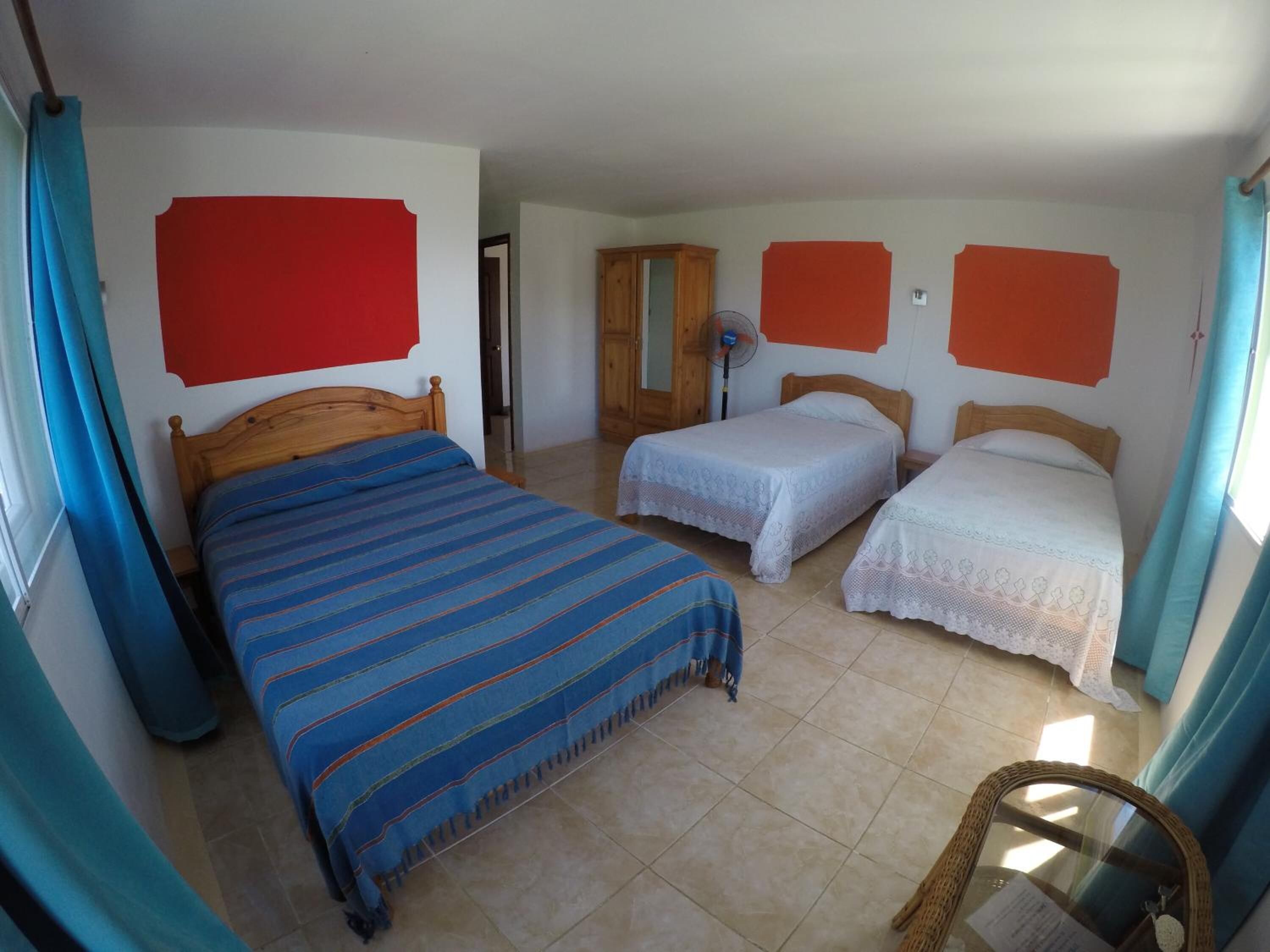 Una habitación con tres camas, un armario de madera, un ventilador y una silla de mimbre.