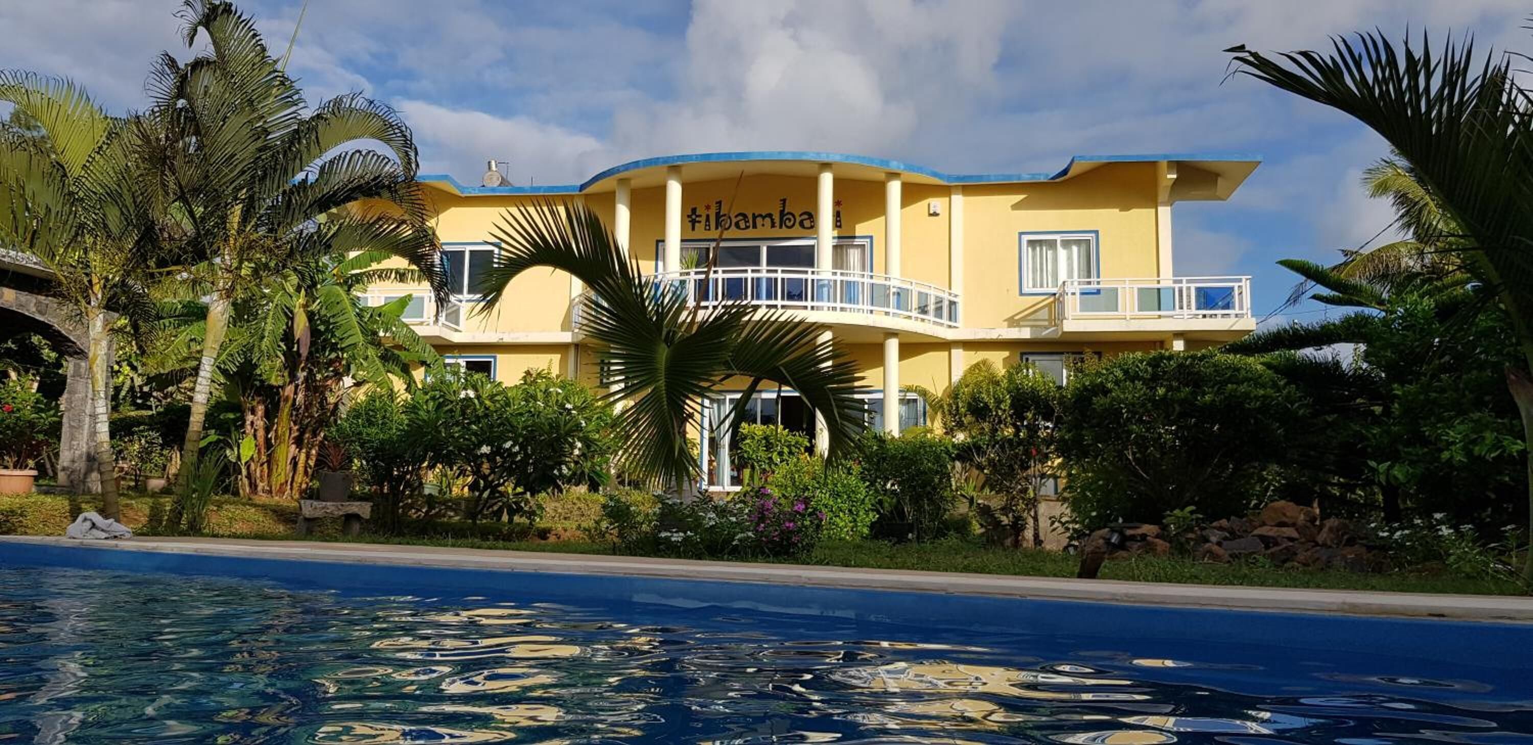Un edificio amarillo con balcones, rodeado de palmeras y una piscina.