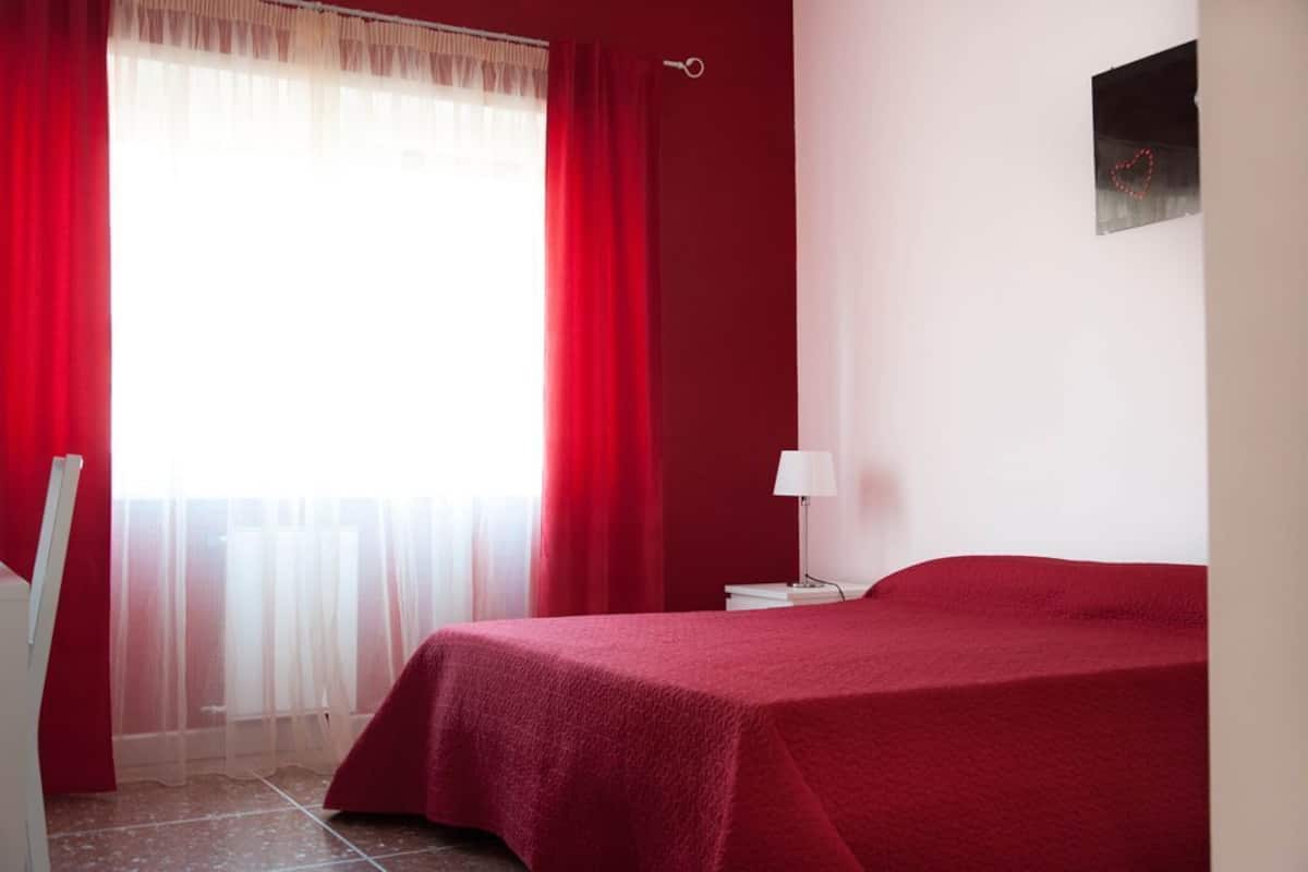 Quarto com cama, mesa de cabeceira, luminária, janela com cortinas leves e quadro na parede.