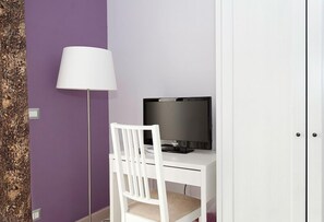 Una scrivania bianca con una televisione, una sedia bianca e una lampada da terra, tutto su uno sfondo viola.