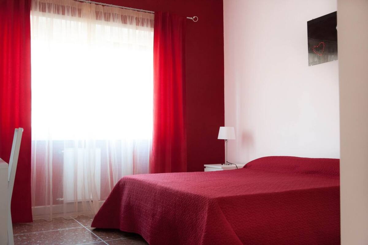 Quarto com cama, mesa de cabeceira, abajur e janela com cortinas vermelhas.