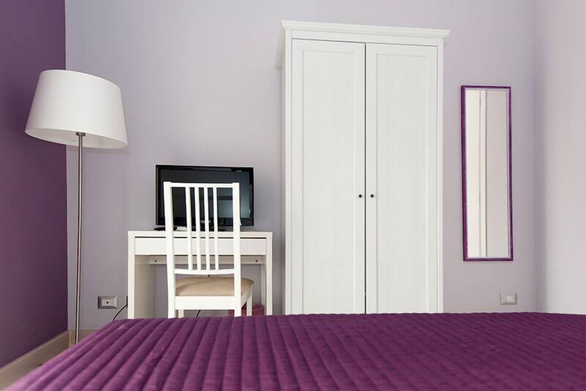Um quarto com cama, escrivaninha com cadeira, televisão e guarda-roupa.