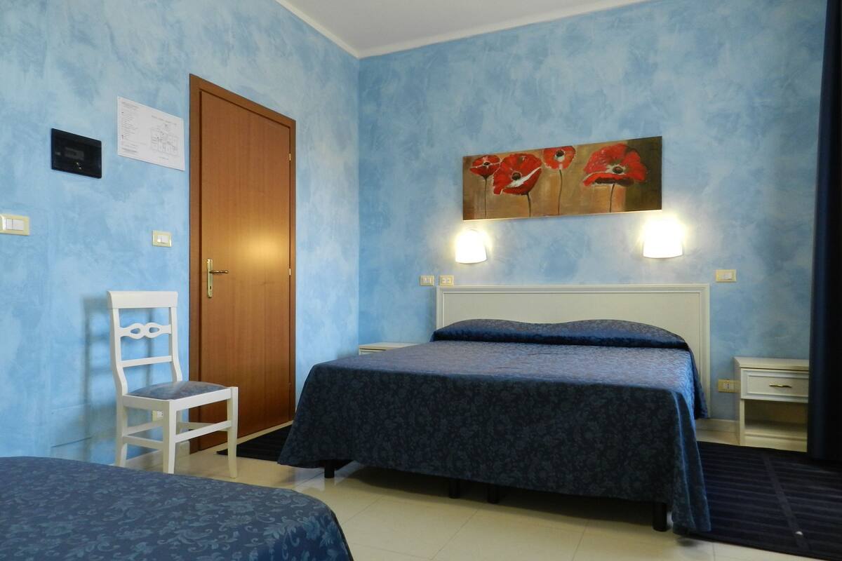 A hotel room with two beds, a wooden door, a white chair, a painting, and a nightstand.