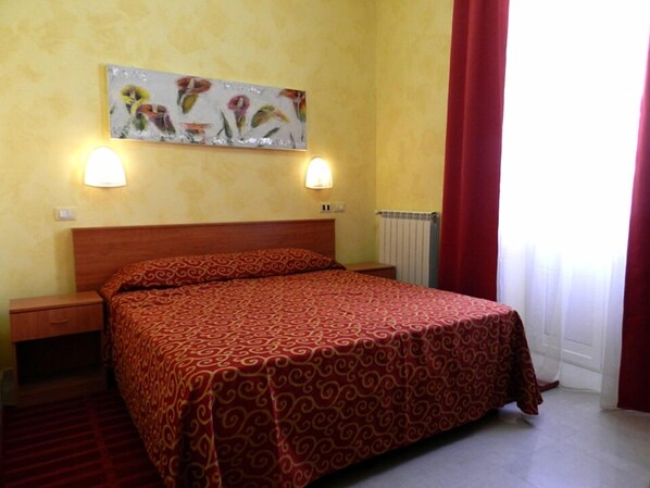 Une chambre d’hôtel comprenant un lit, deux tables de chevet, un luminaire fixé au mur, un radiateur et une fenêtre avec des rideaux.