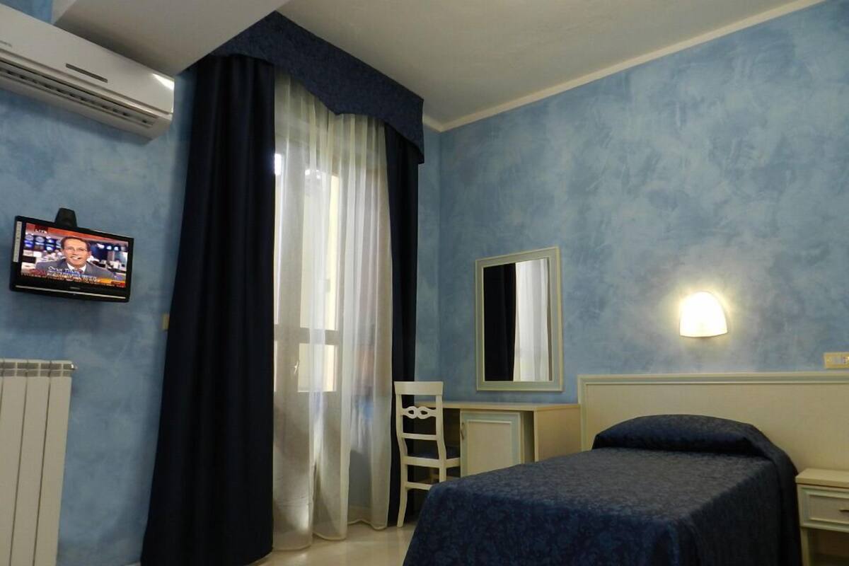 A hotel room with a bed, a bedside table, a mirror, a TV on the wall, and a window with curtains.