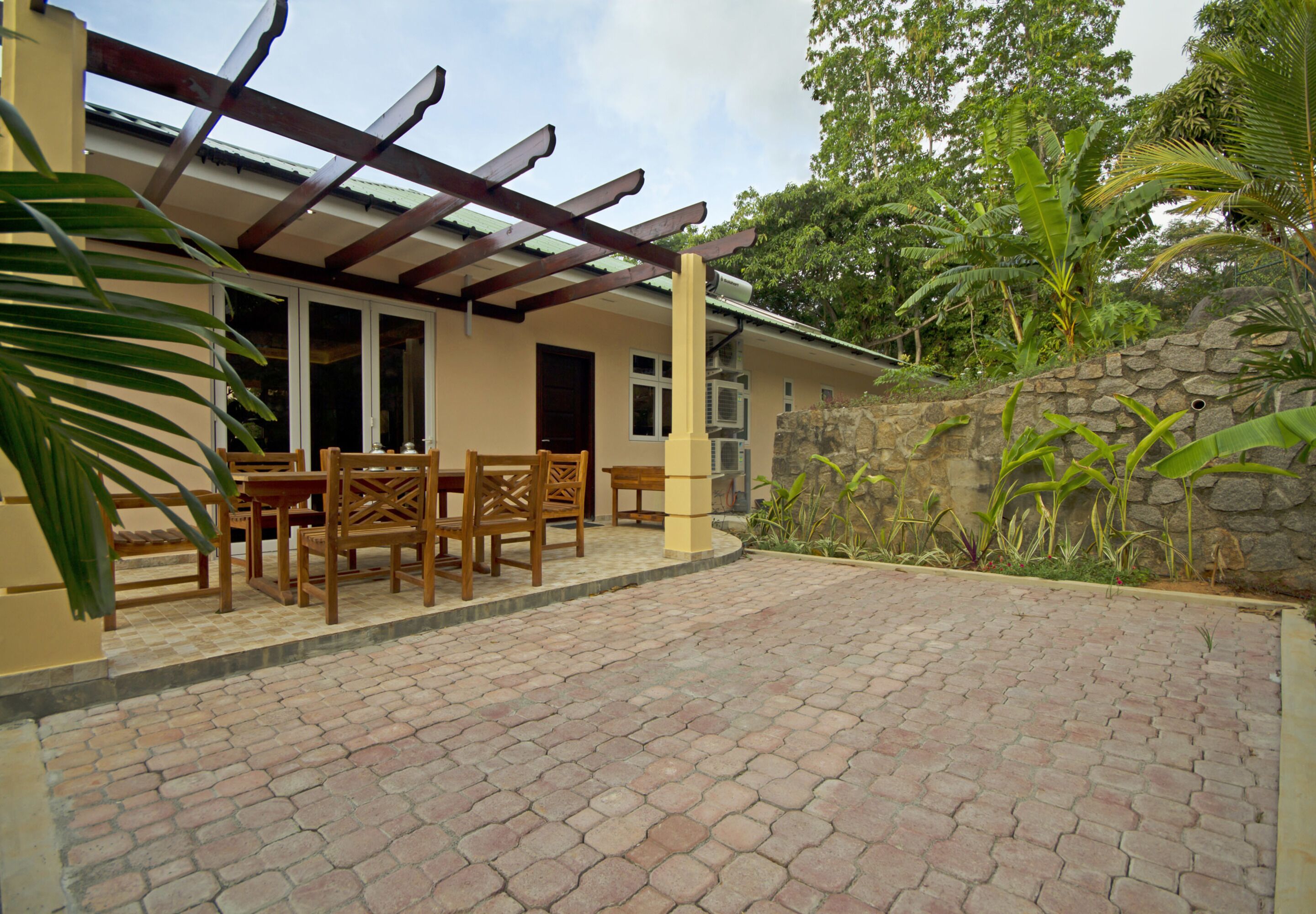 A covered patio with a brick floor, wooden furniture, and a stone wall.