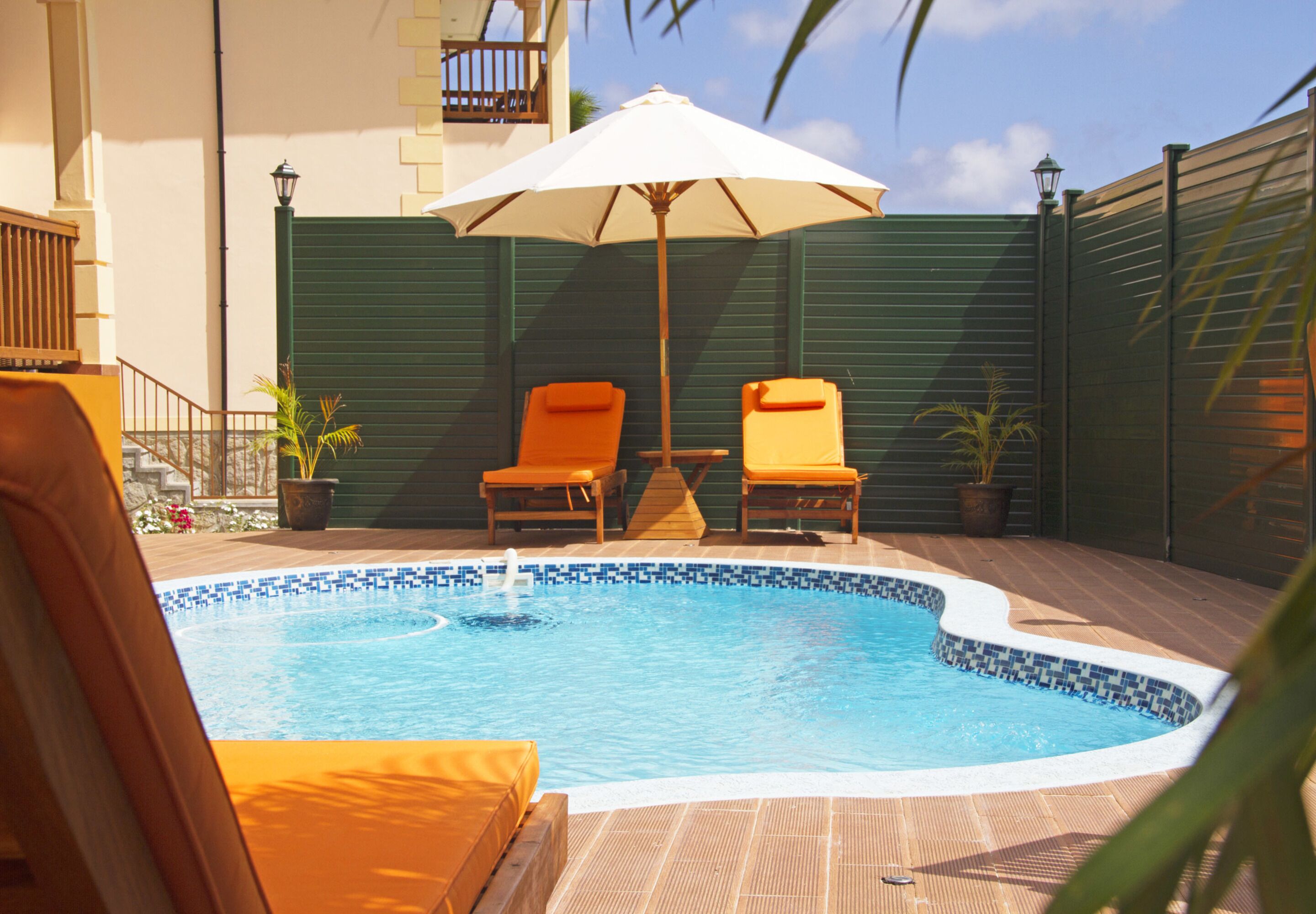 Una piscina con dos sillas reclinables y una sombrilla, rodeada por una terraza de madera y una cerca verde.