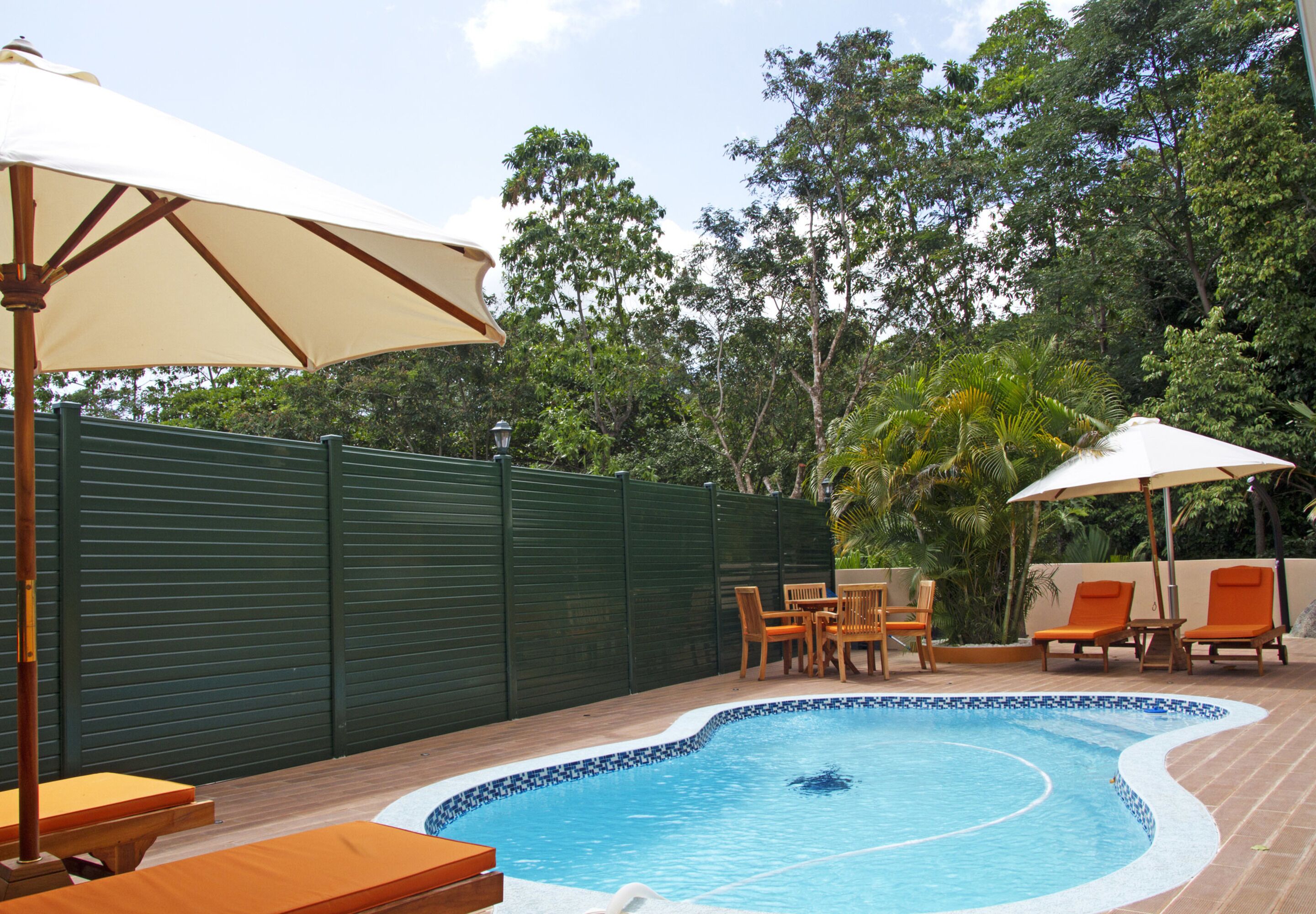 A swimming pool with a tiled edge, surrounded by wooden deck furniture and two umbrellas.