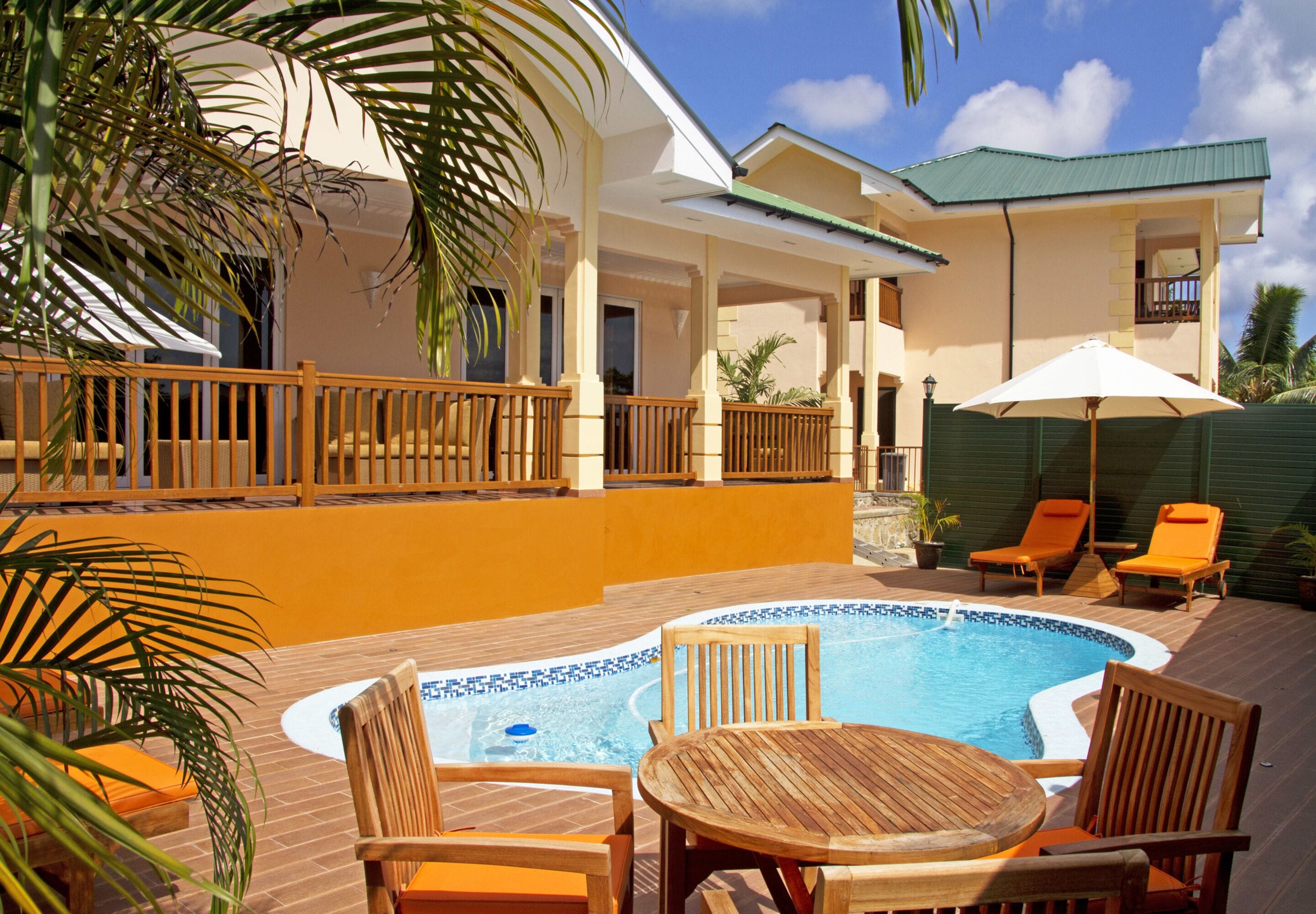 A pool area with wooden chairs and a table, surrounded by a wooden deck and a building with a green roof.