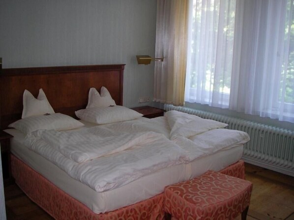 A hotel room with a large bed, a bedside table, a window with sheer curtains, and a radiator.