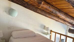 Un dormitorio con techo de madera, cama con ropa de cama blanca, lámpara de noche y cafetera sobre el suelo.