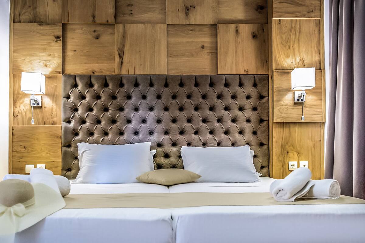 Une chambre d’hôtel avec une tête de lit capitonnée, deux lampes de chevet, un lit avec du linge de lit blanc et une serviette roulée posée sur le lit.