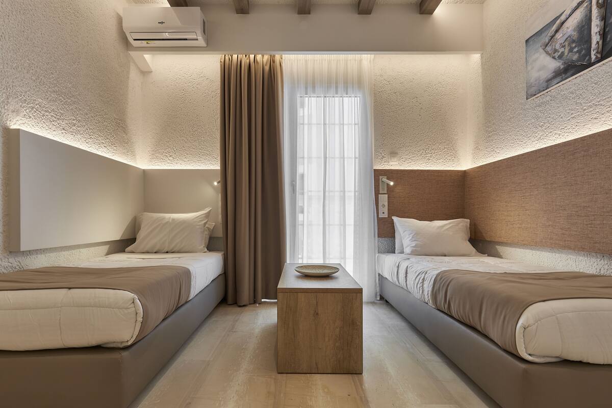 Une chambre d’hôtel moderne avec deux lits, une table de chevet en bois et une grande fenêtre agrémentée de rideaux légers.