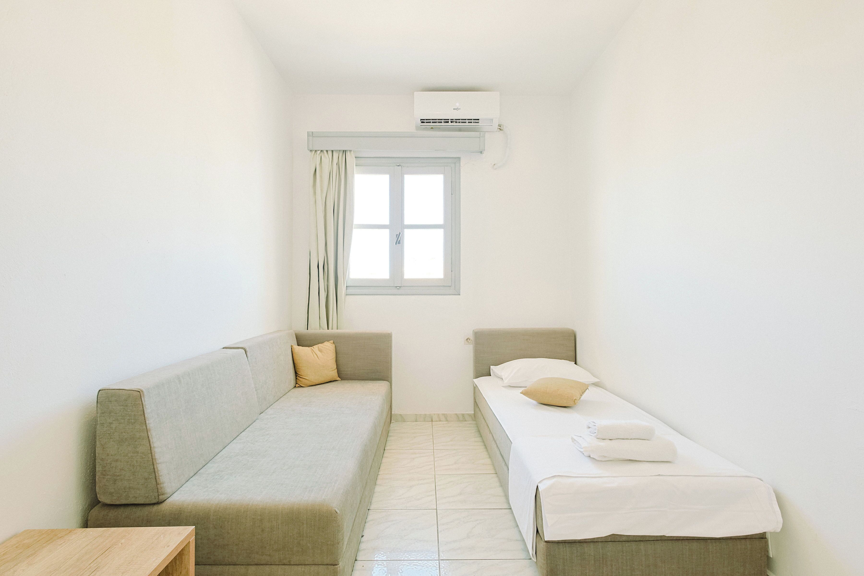 A small, clean room with a bed, a sofa, and an air conditioning unit.