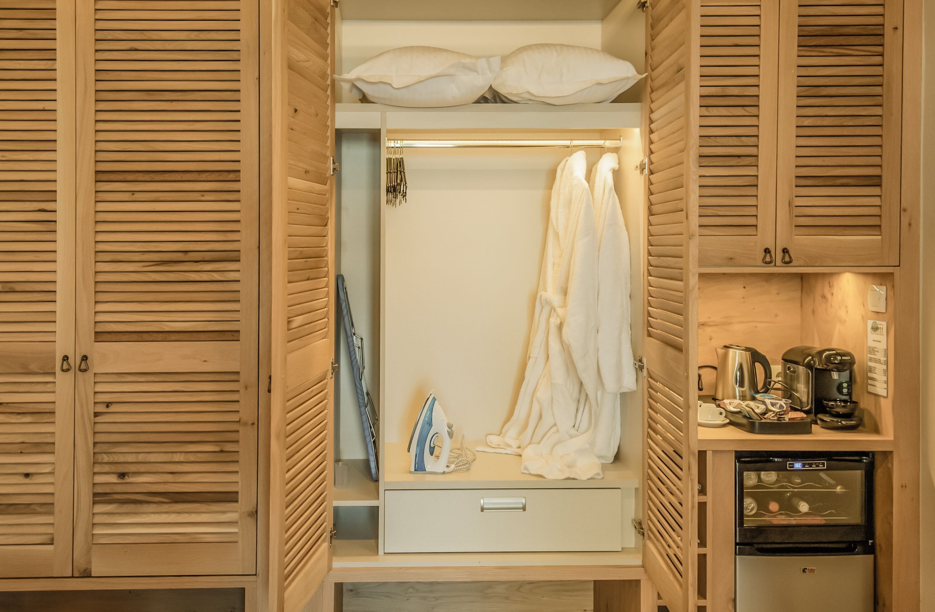 A wooden wardrobe with a hanging robe, an ironing board, and a coffee maker.