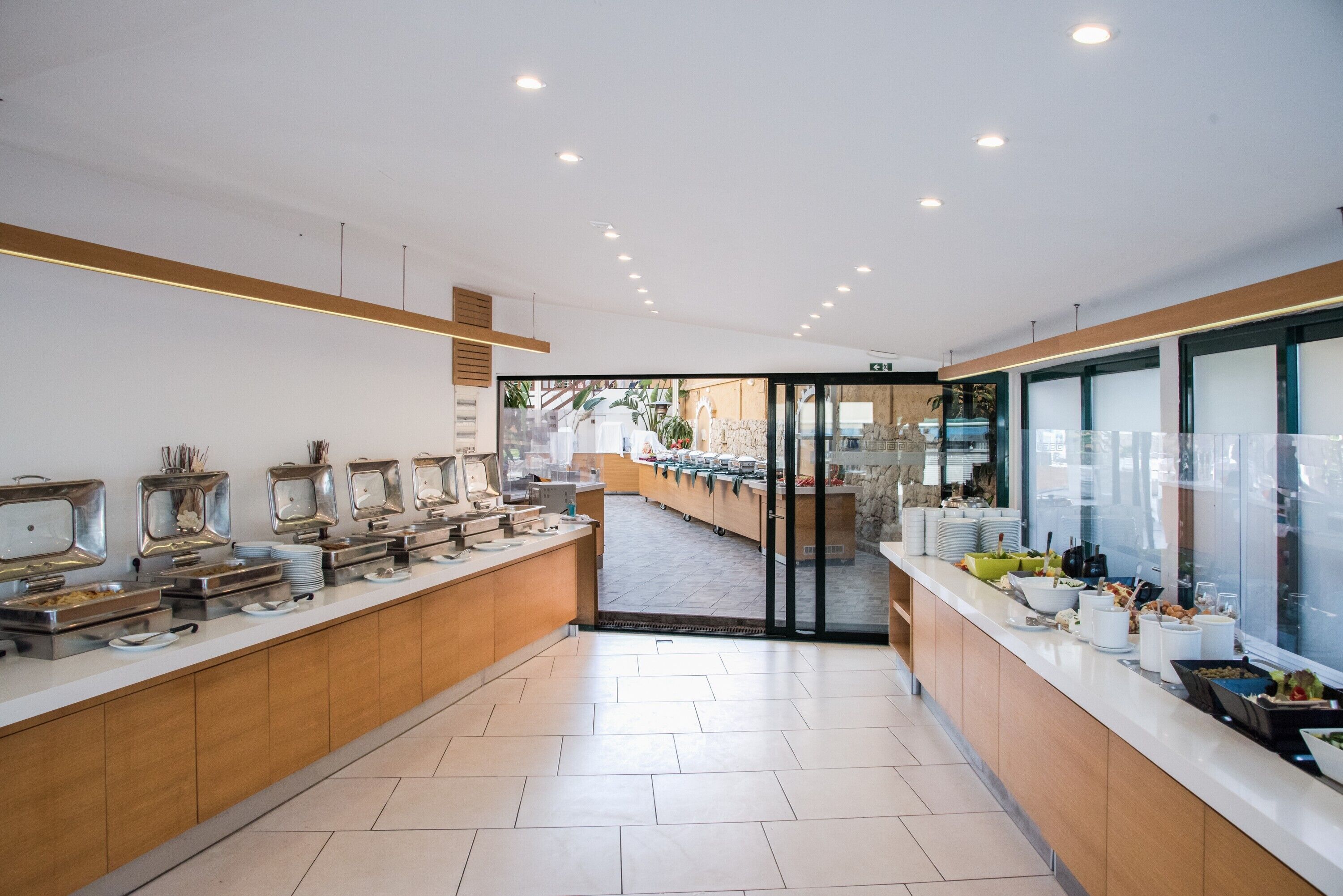 A modern hotel buffet with a long counter, various food stations, and a view of the outdoor area through a glass door.