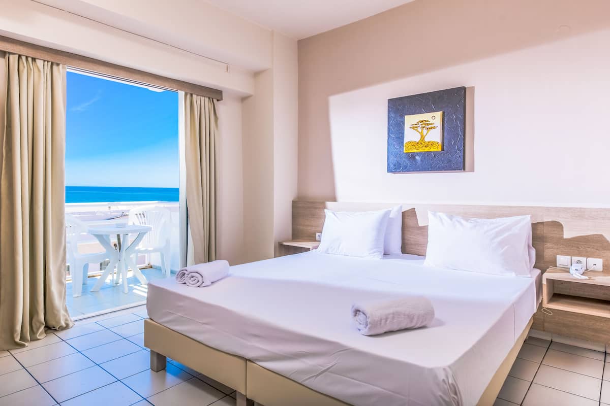 Une chambre d’hôtel avec un grand lit, une vue sur la mer, un balcon avec une table et des chaises, et un tableau encadré au mur.