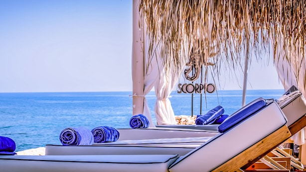 Eine Strandlounge mit Liegestühlen, einem Reetdach und Blick auf das Meer.