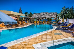 A pool area with lounge chairs and umbrellas, a thatched-roof structure, and a view of the sea. - Porto Greco Village Beach Hotel - All Inclusive (Hersonissos)