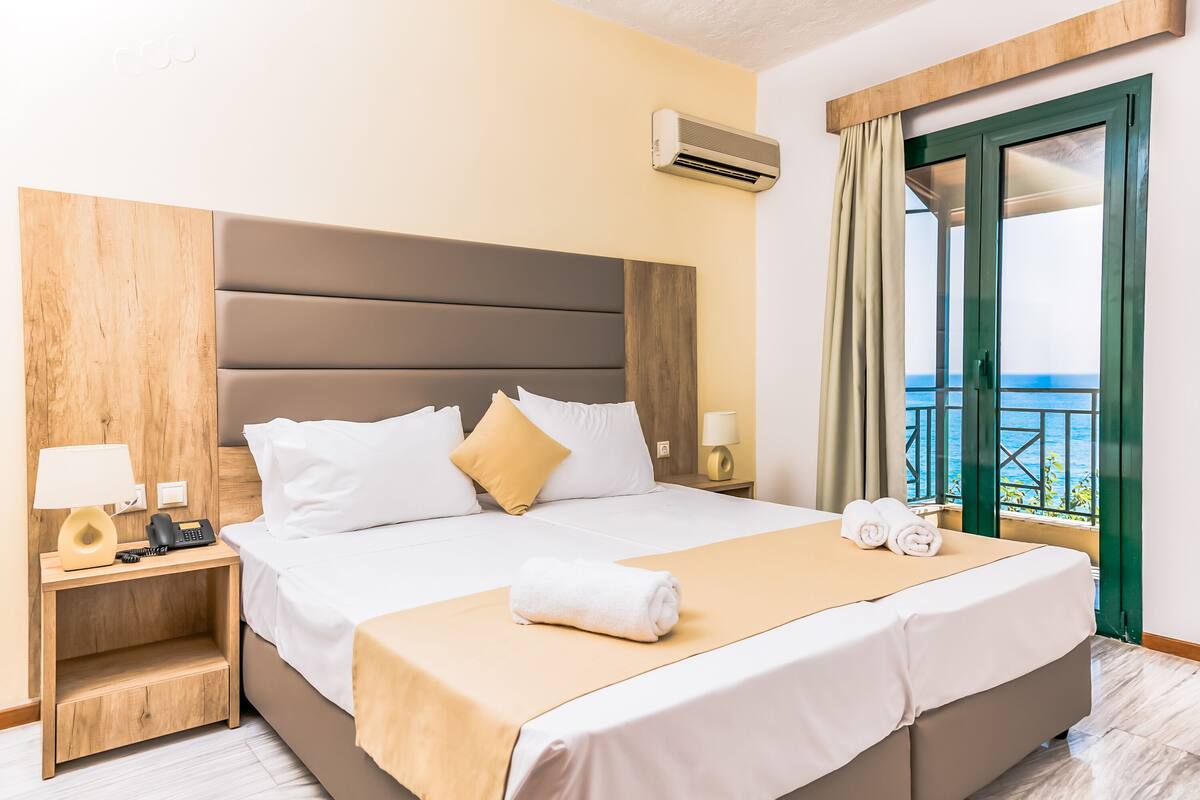 Une chambre d’hôtel avec un lit, une table de chevet avec une lampe, un téléphone et une vue sur la mer depuis la porte-fenêtre donnant sur le balcon.