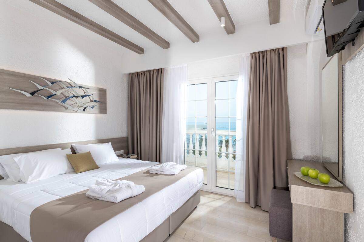 Une chambre d’hôtel avec un lit, un balcon avec des rideaux, un téléviseur fixé au mur et une petite table avec des pommes.