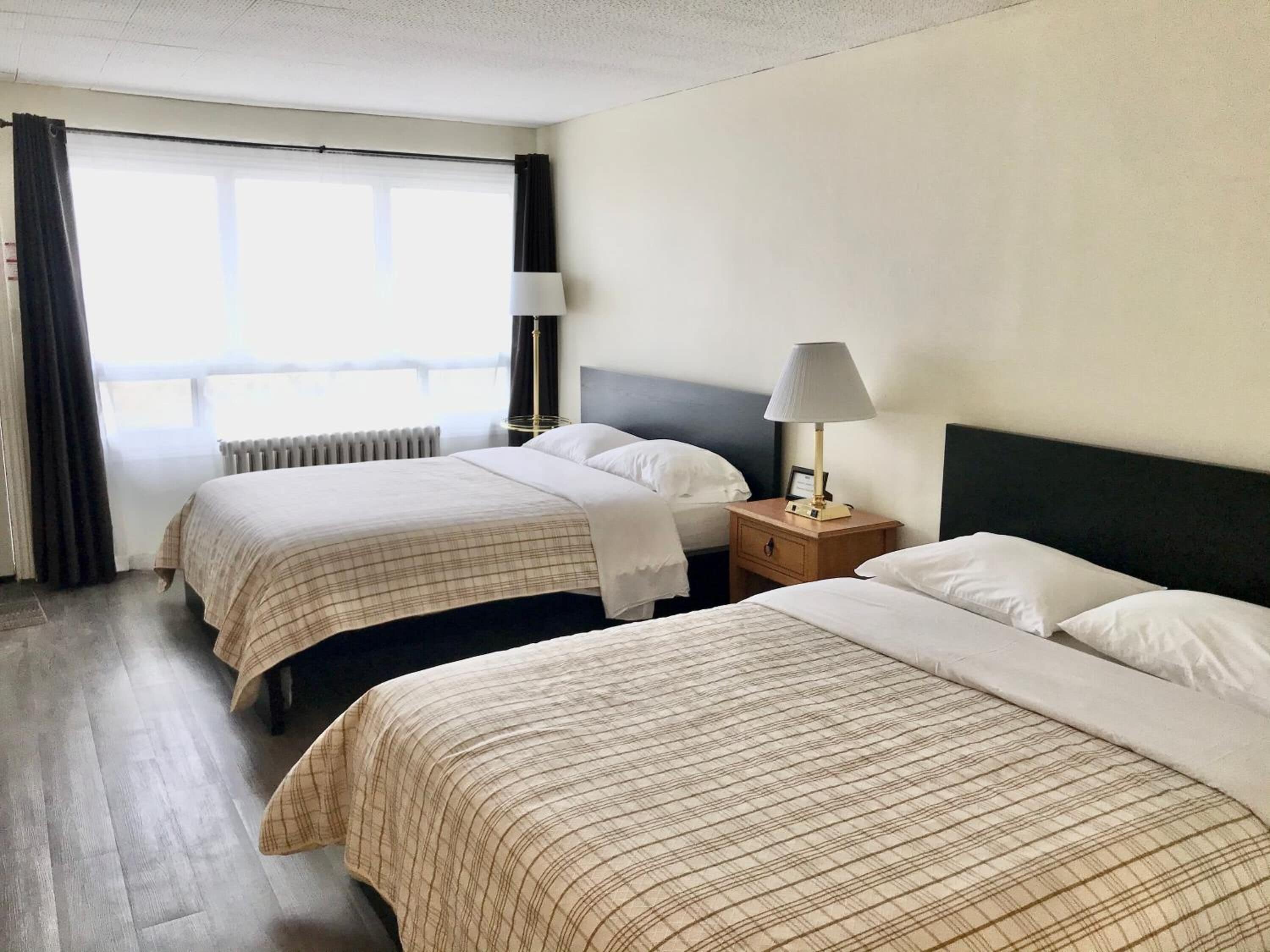 A hotel room with two beds, a window with curtains, a radiator, and a nightstand with a lamp.