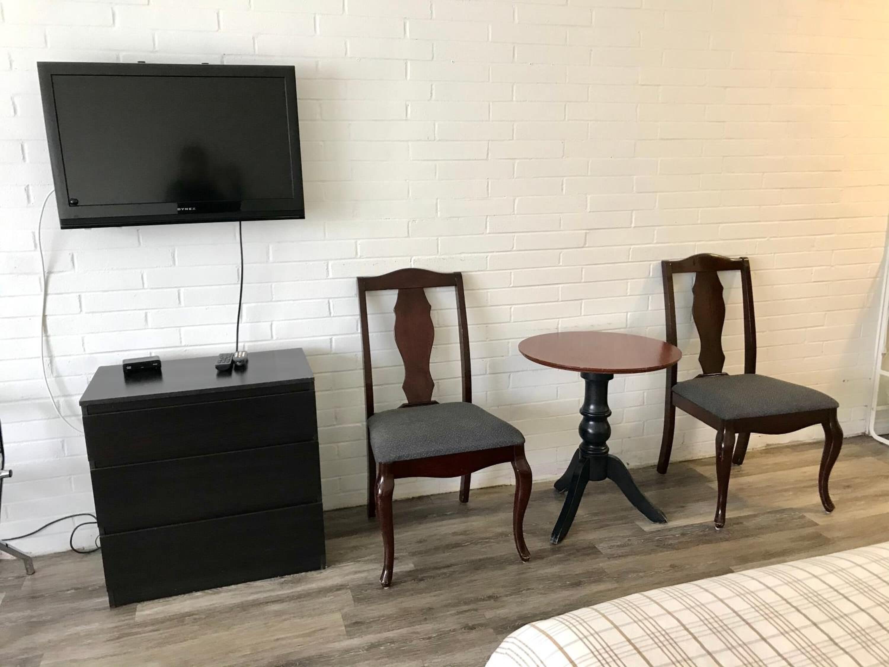 A room with a bed, a TV mounted on the wall, a small wooden table, two chairs, and a dresser.