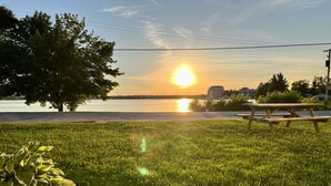 A grassy area with a wooden picnic table and a tree, overlooking a body of water with buildings in the distance. - Lakeside Inn (Ramara)