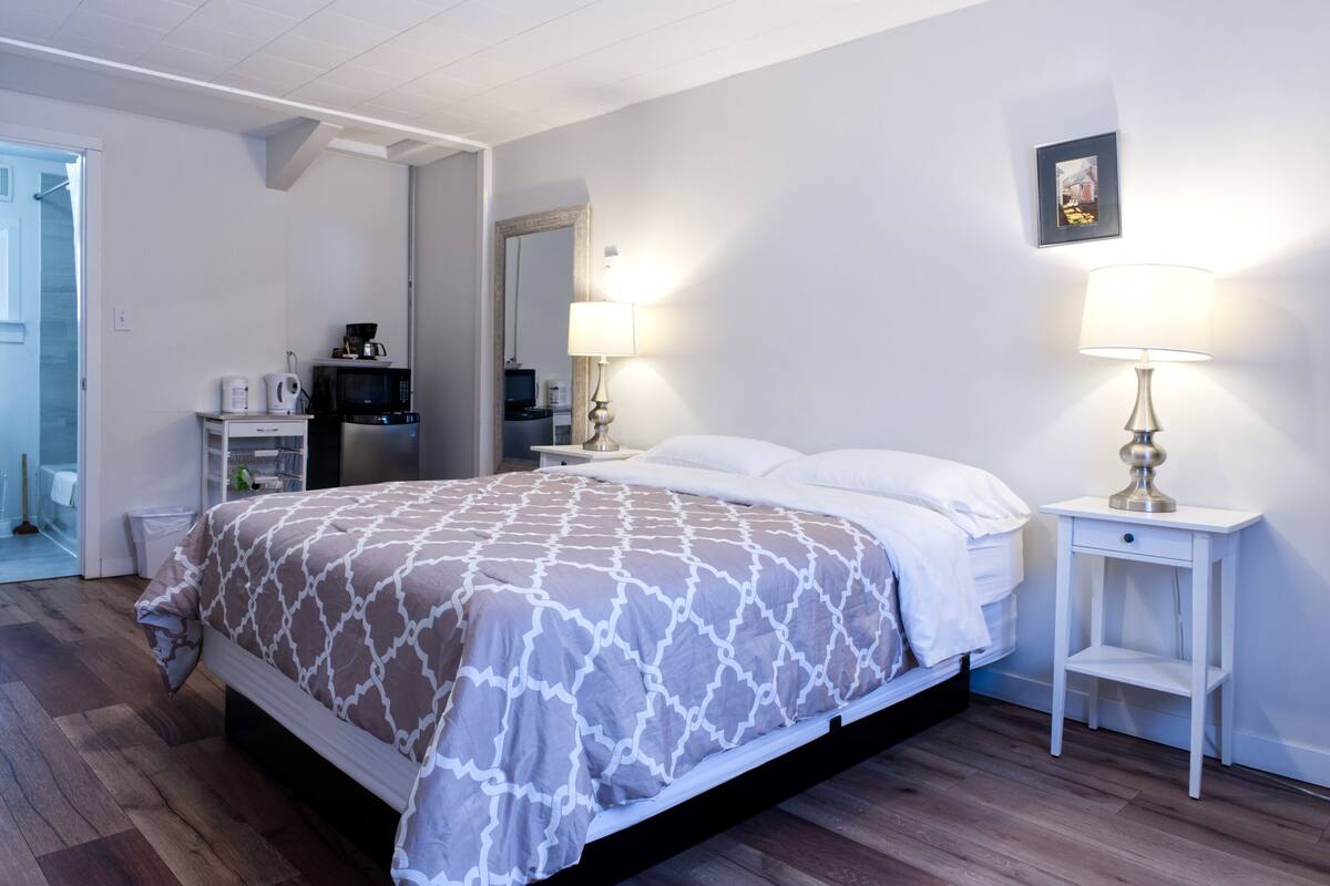 A bedroom with a bed, bedside tables, a lamp, a mirror, and a kitchenette area.