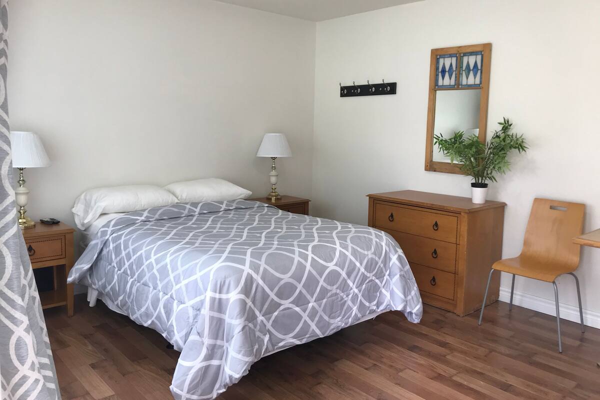 A bedroom with a bed, two bedside tables with lamps, a wooden dresser, a chair, a mirror, and a potted plant.