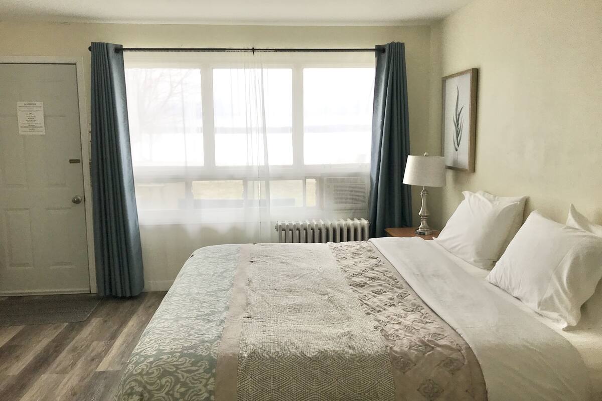 A hotel room with a large bed, a radiator, a nightstand with a lamp, and a window with curtains.