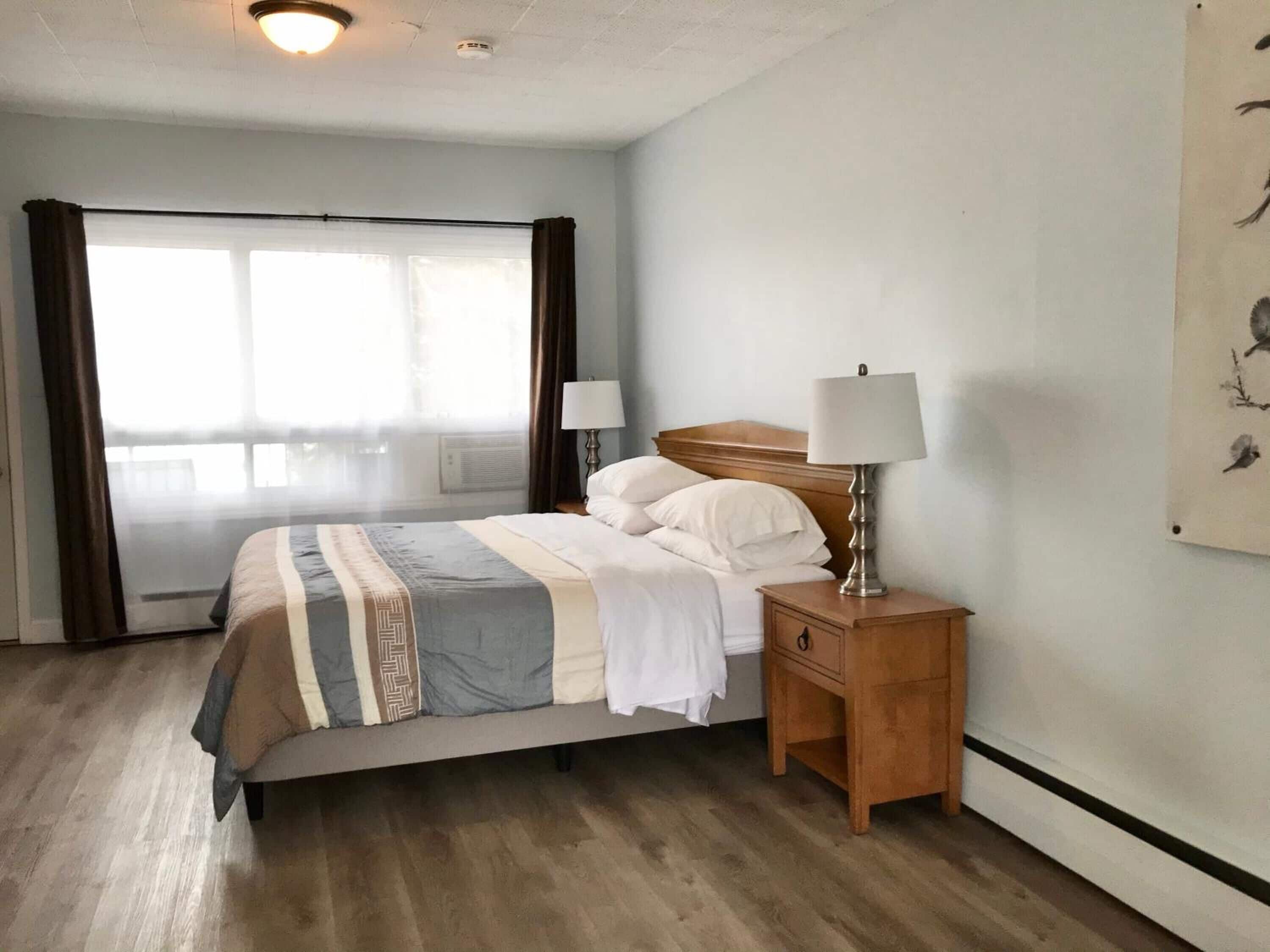 A hotel room with a bed, a nightstand with a lamp, a window with curtains, and a wall with a framed picture.