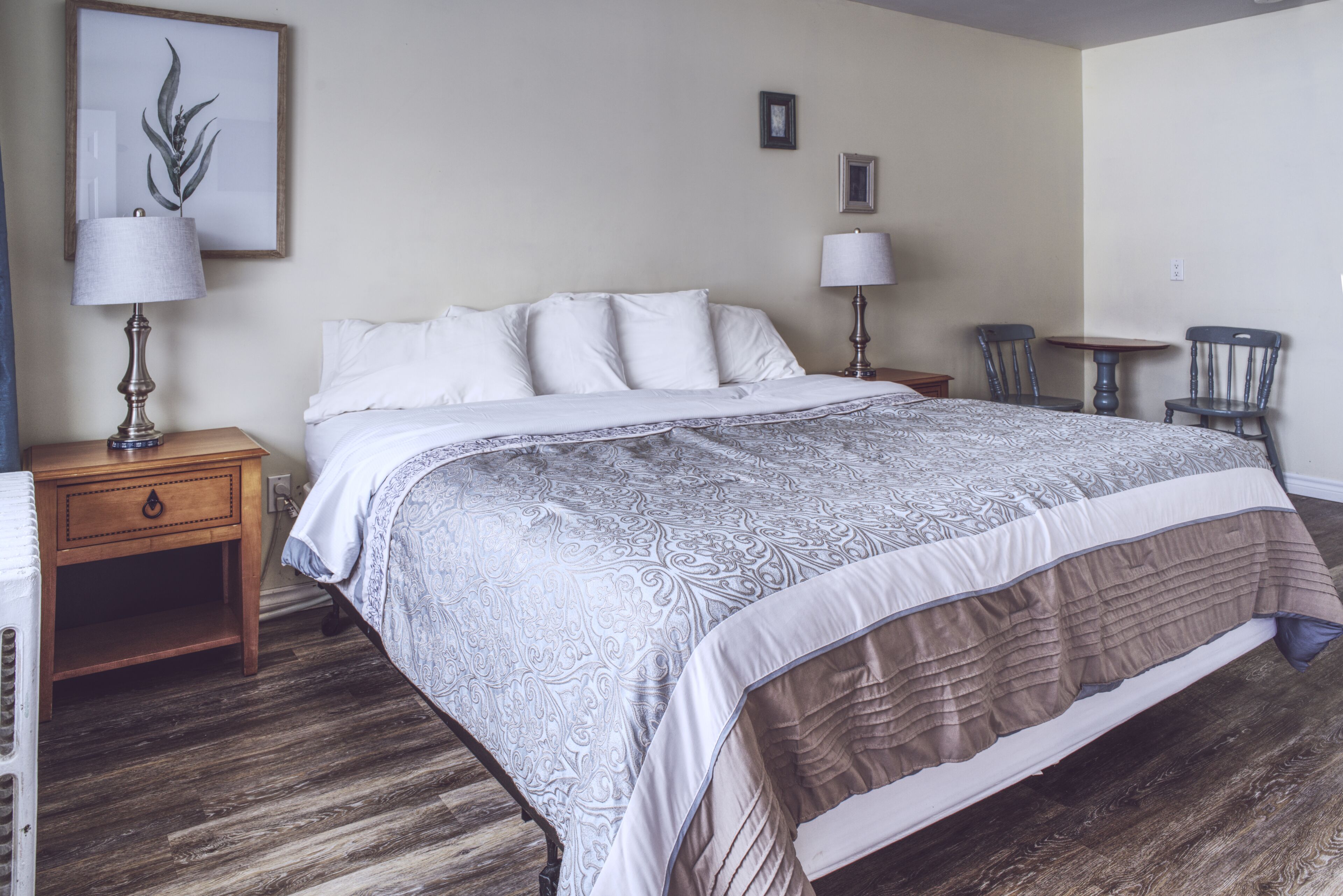 A neatly arranged bedroom with a large bed, two bedside tables with lamps, a small table with chairs, and a framed picture on the wall.