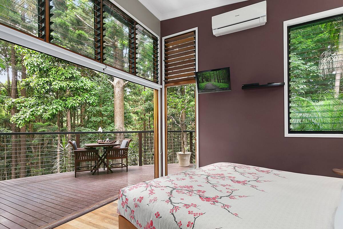 A bedroom with a large window, a bed with a floral bedspread, a TV on the wall, and a balcony with a table and chairs.