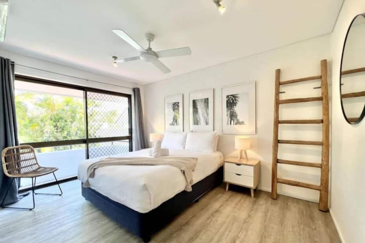 Une chambre moderne comprenant un lit, une chaise, une table de chevet, un ventilateur de plafond et une fenêtre donnant sur un paysage verdoyant.