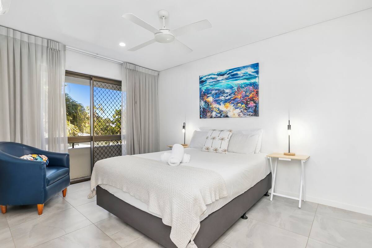 Une chambre à coucher comprenant un lit, une chaise, une table de chevet, un ventilateur de plafond et une grande fenêtre avec des rideaux.