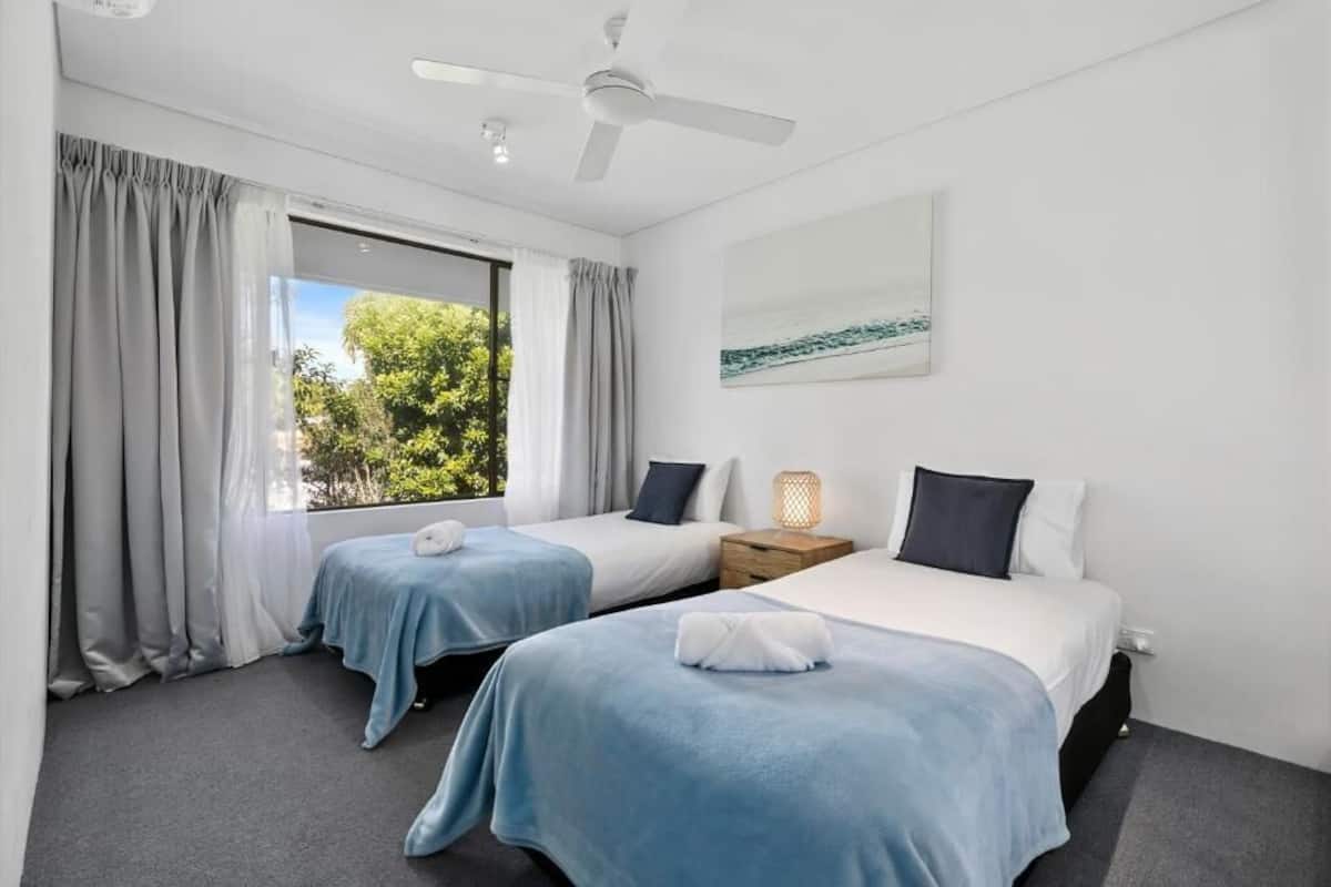 Une chambre d’hôtel avec deux lits simples, un ventilateur de plafond, une fenêtre avec des rideaux, une table de chevet avec une lampe et un tableau au mur.