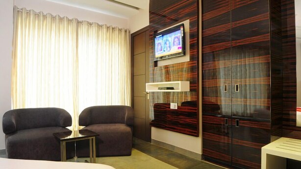 A hotel room with a large bed, a TV mounted on the wall, a seating area with two chairs, and a wardrobe with a mirror. - Hotel Gulnar (New Delhi)