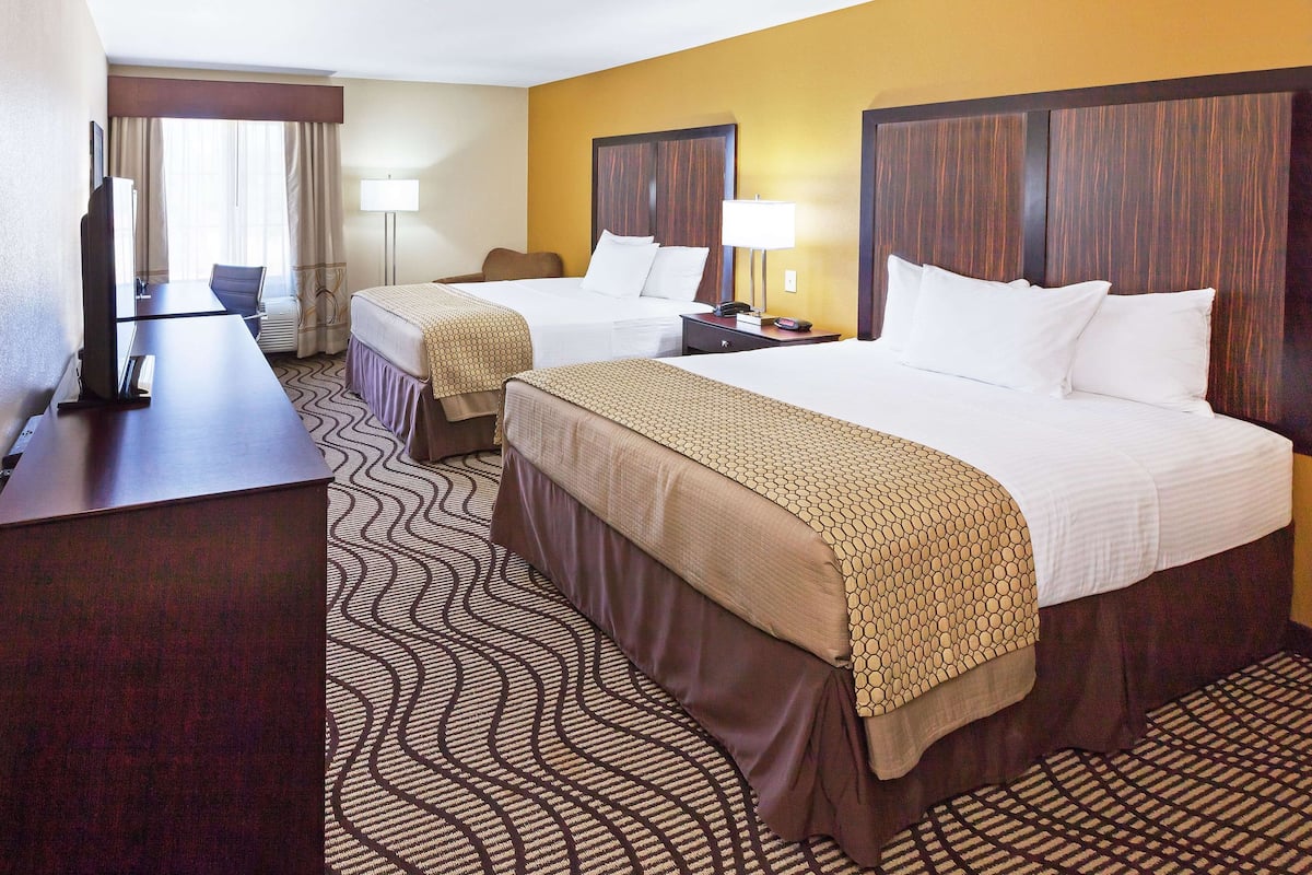 A hotel room with two beds, a desk, a chair, a TV, and a lamp.