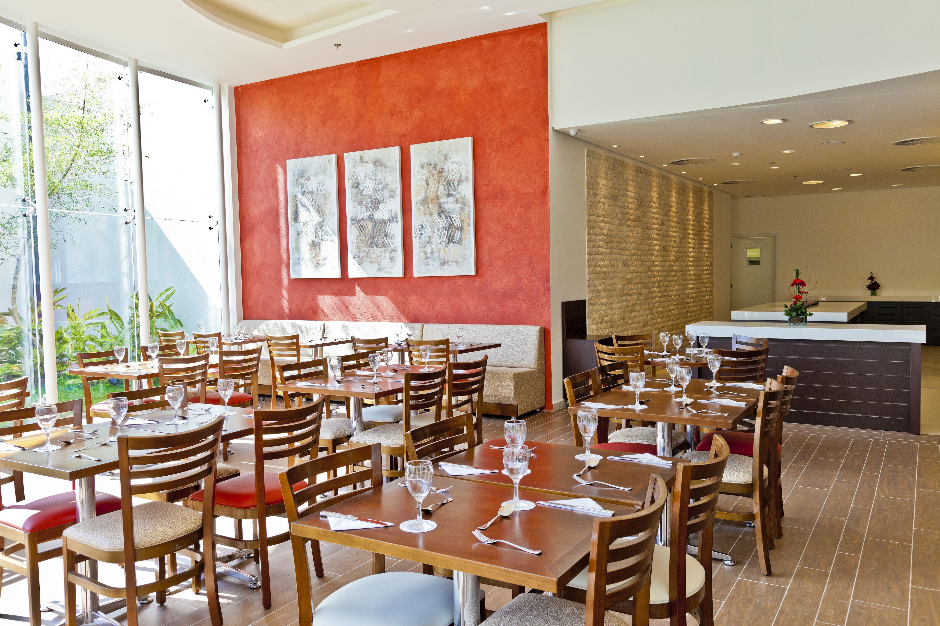 Un restaurante con varias mesas y sillas, un muro rojo decorado con obras de arte, y vistas a zonas verdes al exterior.