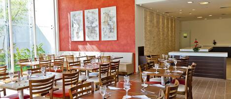 Un restaurante con varias mesas y sillas, un muro rojo decorado con obras de arte, y vistas a zonas verdes al exterior.